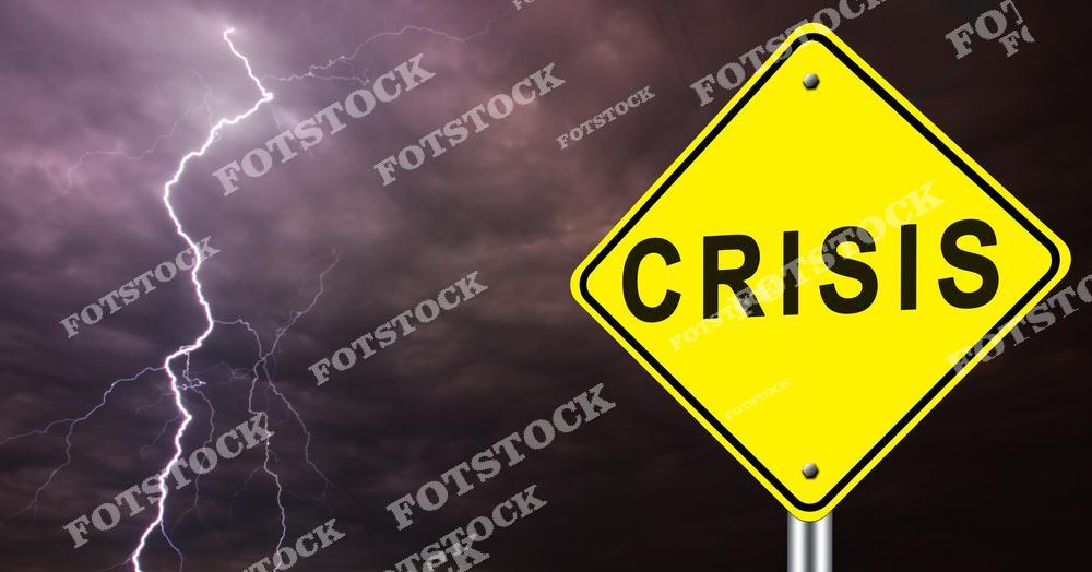 На изображении желтый дорожный знак с надписью "Crisis", установленный на фоне темного грозового неба с молнией. Контраст яркого знака и мрачного фона подчеркивает драматизм ситуации. Фотография символизирует предупреждение о кризисе, экономических или социальных потрясениях.